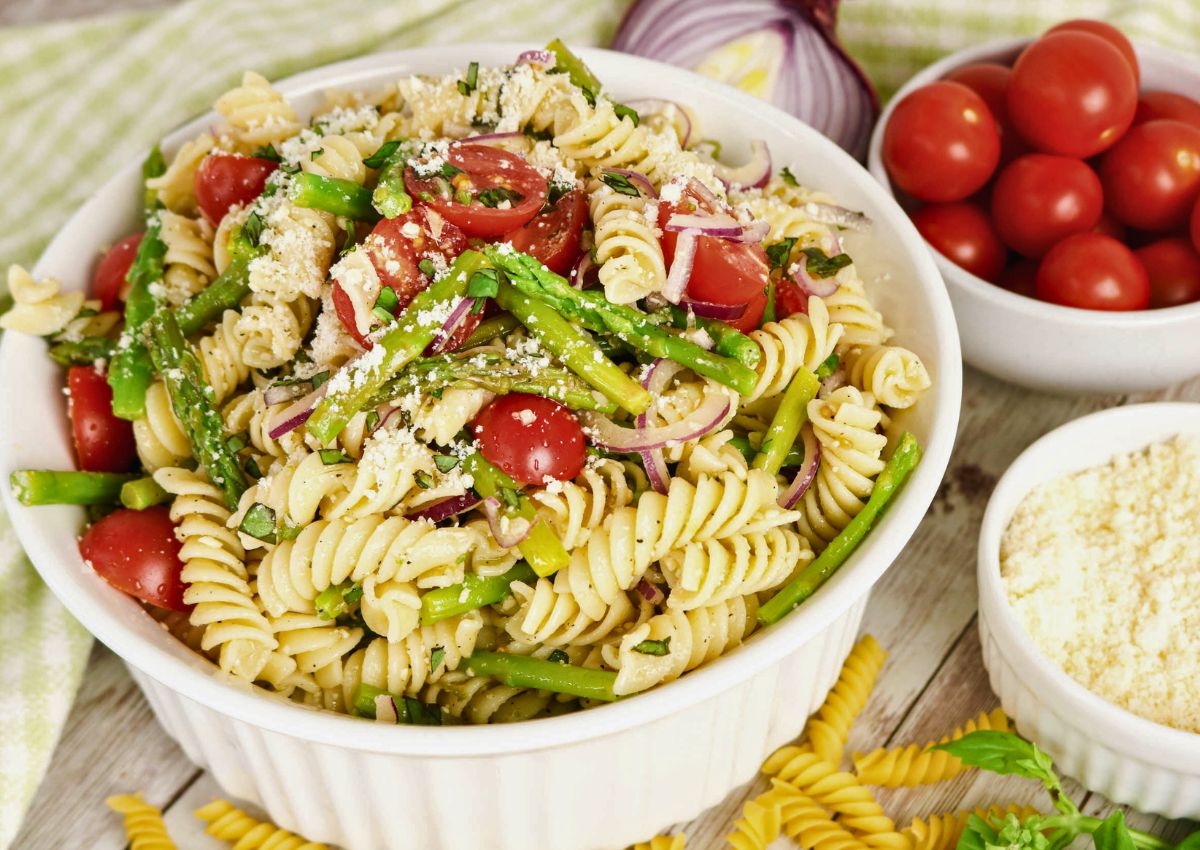 A bowl of rotini pasta salad with asparagus, cherry tomatoes, red onion, and grated cheese, surrounded by small bowls of cherry tomatoes and grated cheese.