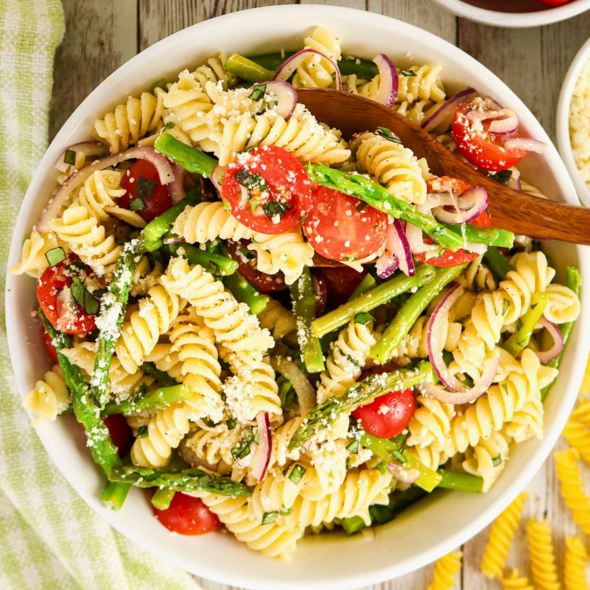 A bowl of rotini pasta salad with asparagus, cherry tomatoes, red onion, and grated cheese, mixed with herbs.