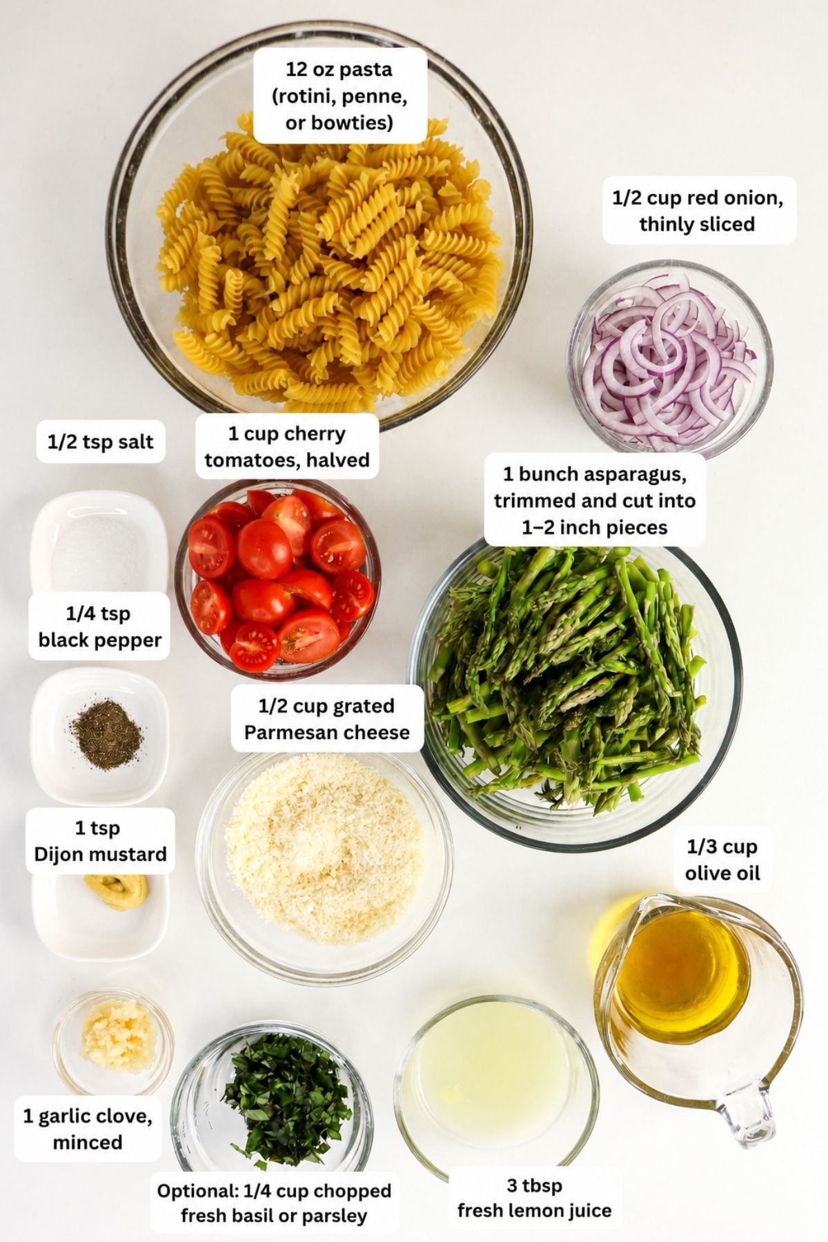 Ingredients for a pasta salad are arranged on a white surface, including pasta, red onion, cherry tomatoes, asparagus, cheese, oil, lemon juice, and seasonings in separate bowls.