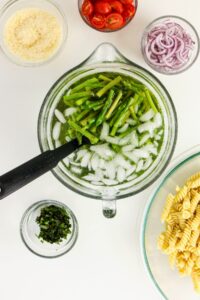 Chopped asparagus in ice water with a slotted spoon, surrounded by bowls of pasta, sliced red onion, cherry tomatoes, breadcrumbs, and chopped herbs.