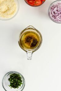 A glass measuring cup with oil and pepper in the center, surrounded by small bowls of grated cheese, sliced tomatoes, red onions, and chopped herbs on a white surface.