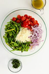 A glass bowl with asparagus, cherry tomatoes, red onion, grated cheese, and herbs; a small bowl of chopped herbs and a cup of dressing are nearby.