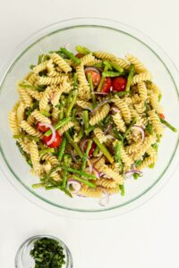 A glass bowl filled with rotini pasta salad containing asparagus, cherry tomatoes, red onion, and herbs. A small bowl of chopped herbs sits beside it.