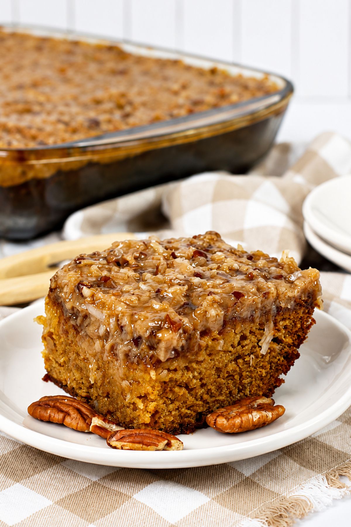 A slice of moist cake topped with a coconut and pecan topping is served on a white plate with pecans, with a larger cake in a glass dish in the background.