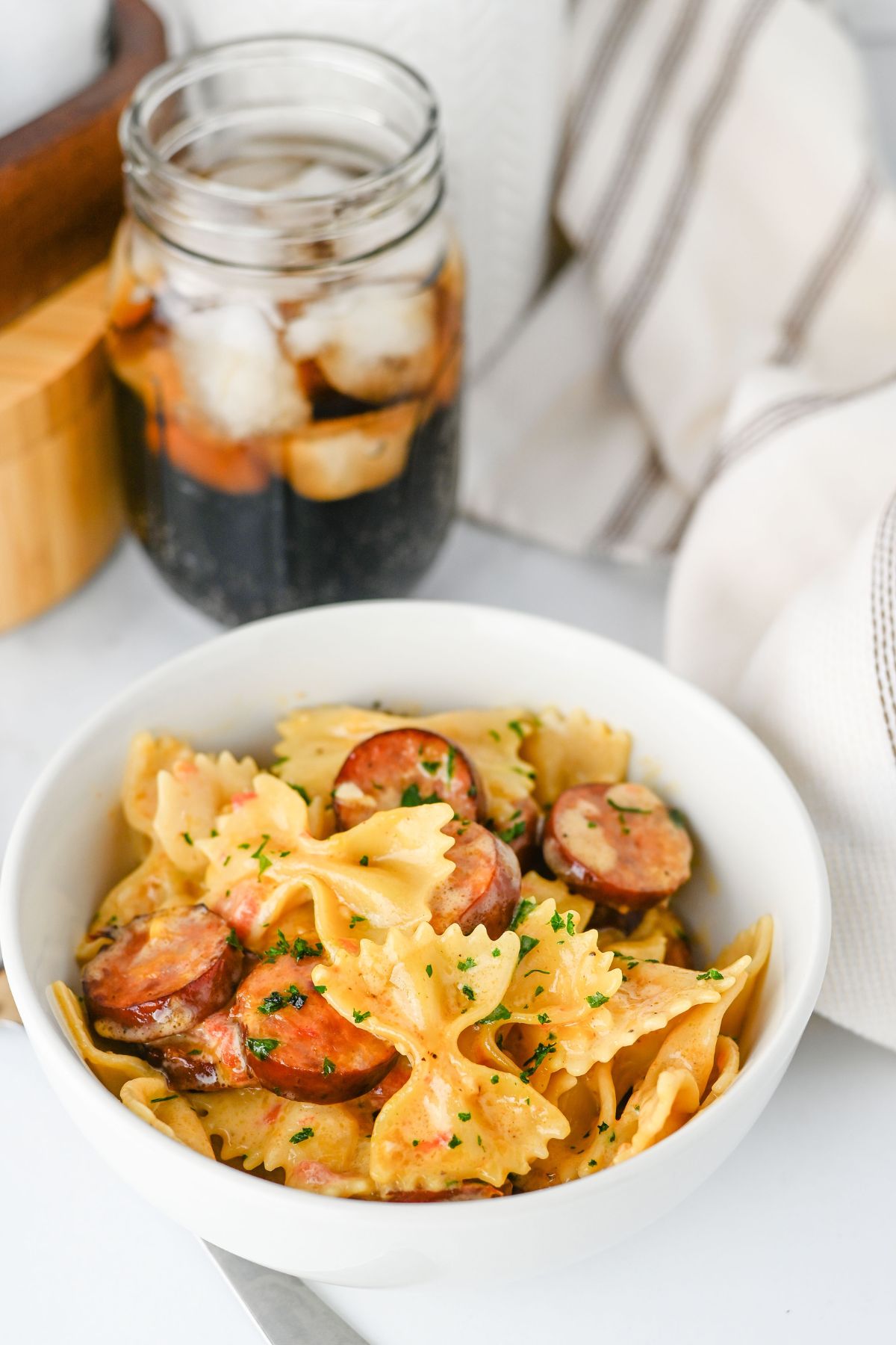 A white bowl of bowtie pasta with sliced sausage and herbs, with a jar of iced dark soda in the background.