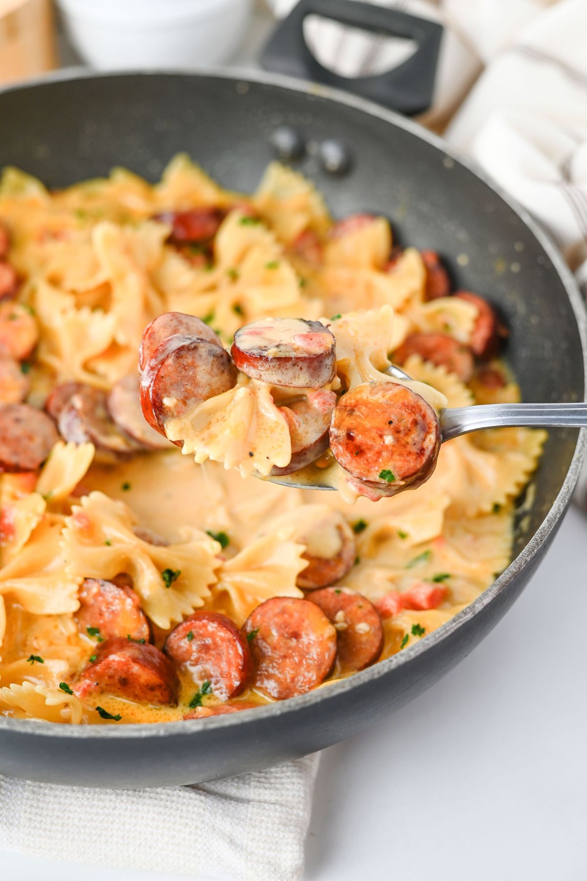 A spoon holds sliced sausage and bowtie pasta in a creamy sauce above a pan filled with the same pasta dish.