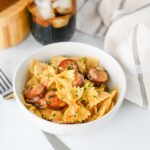 A white bowl filled with bowtie pasta and sliced sausage, garnished with herbs, sits on a white surface next to a drink with ice and a striped napkin.