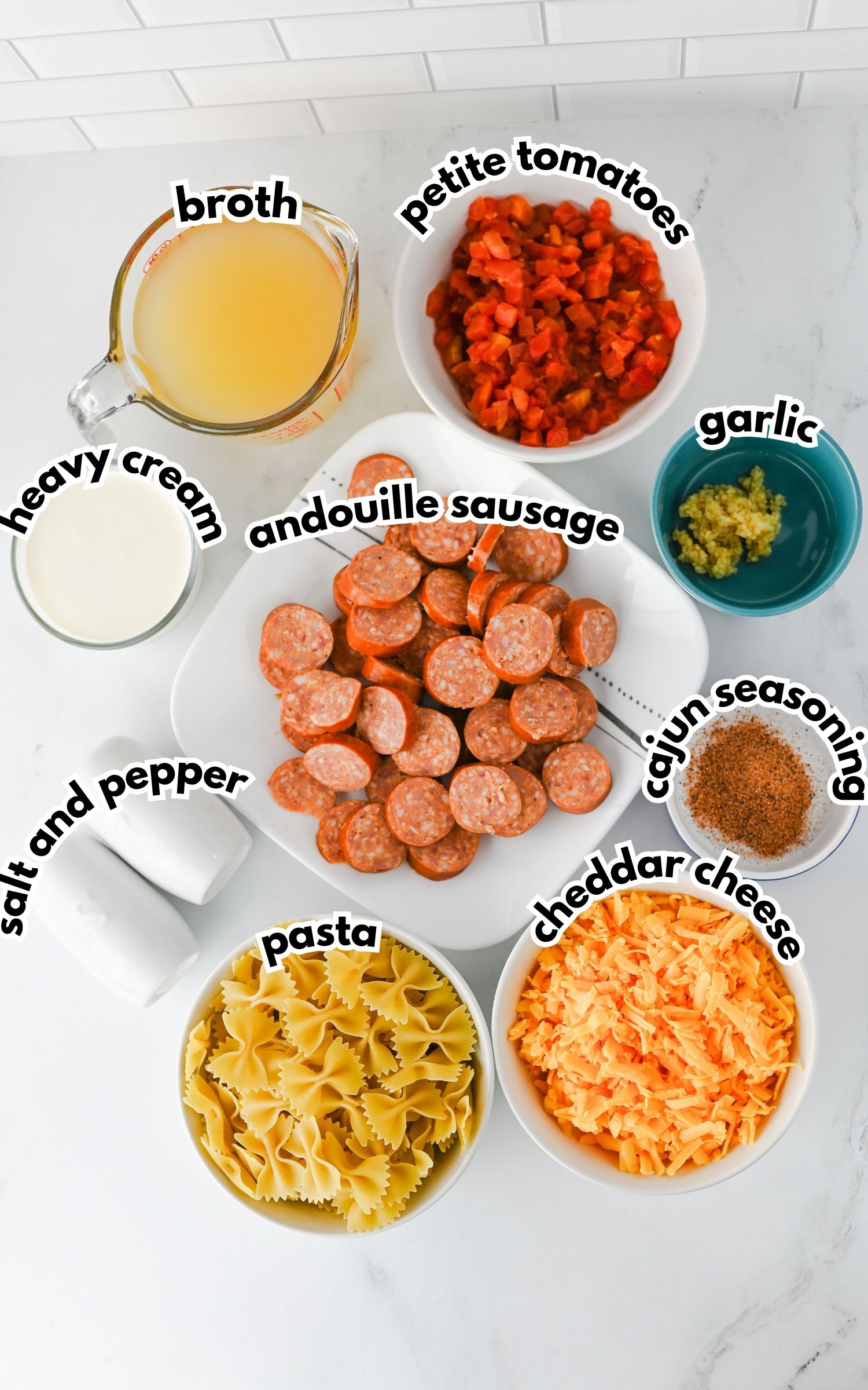 Bowls and containers of pasta, sliced andouille sausage, shredded cheddar cheese, petite tomatoes, heavy cream, broth, minced garlic, Cajun seasoning, salt, and pepper on a countertop.