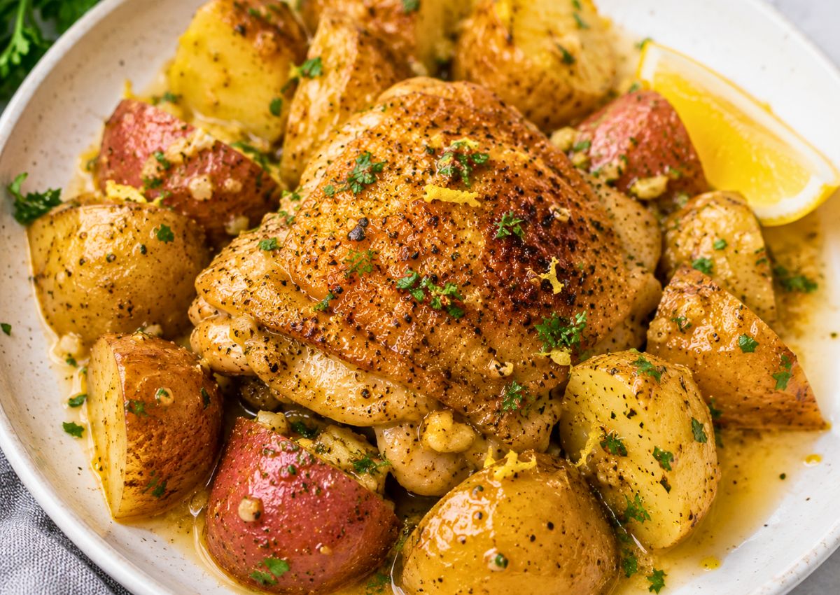 A plate of roasted chicken thigh with seasoned potato wedges, garnished with herbs and lemon wedge.