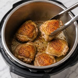 Five seasoned chicken thighs are being browned in an Instant Pot, with metal tongs holding one piece above the rest.