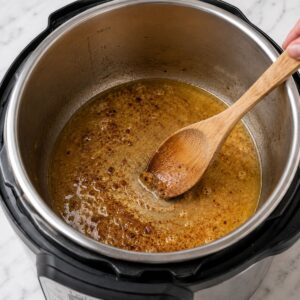 A wooden spoon stirs a brown sauce in an open Instant Pot on a marble countertop.