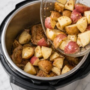 Chopped seasoned potatoes are being added to an instant pot containing seasoned chicken thighs.
