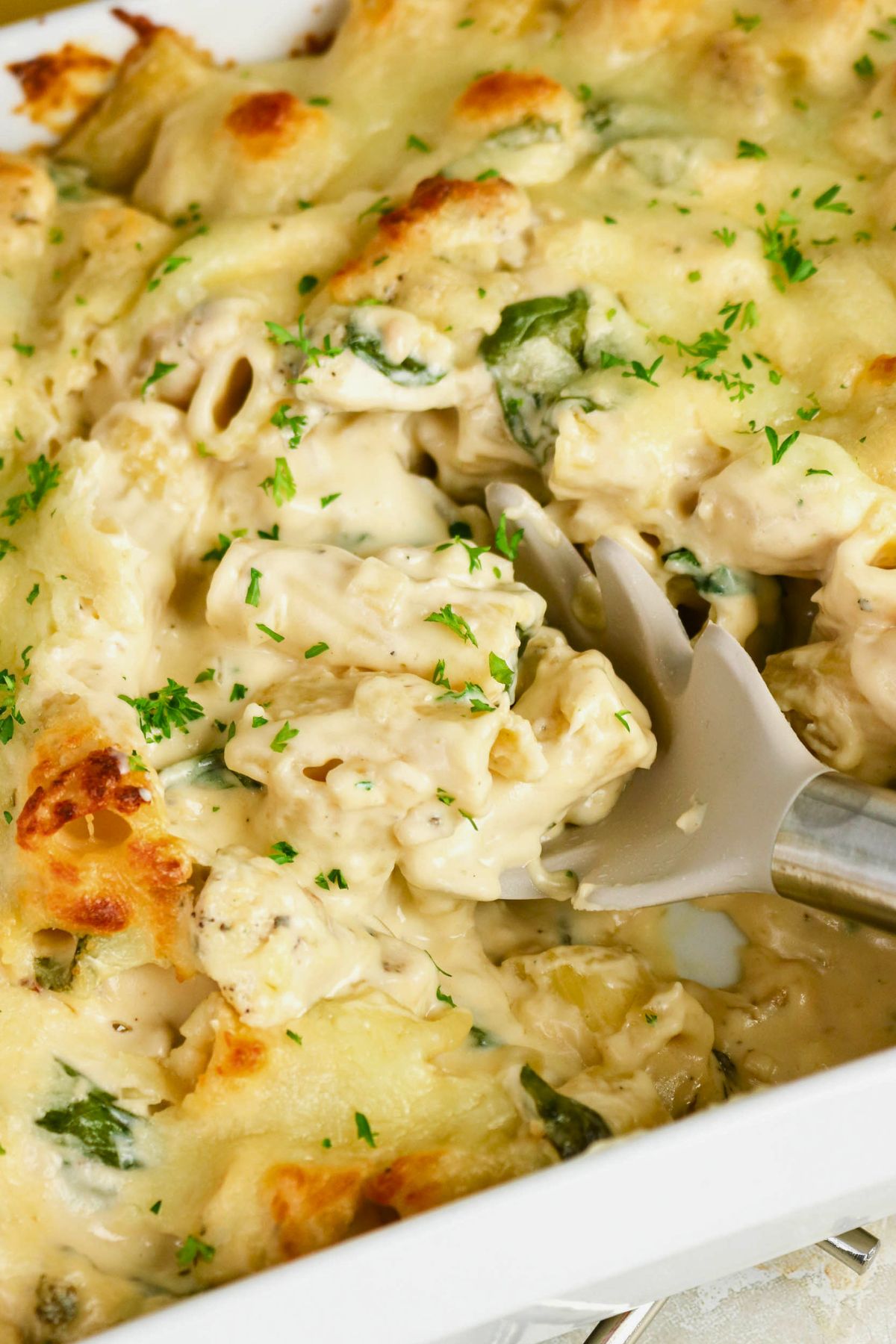 A serving spoon scoops creamy chicken and spinach casserole topped with melted cheese and chopped herbs from a white baking dish.