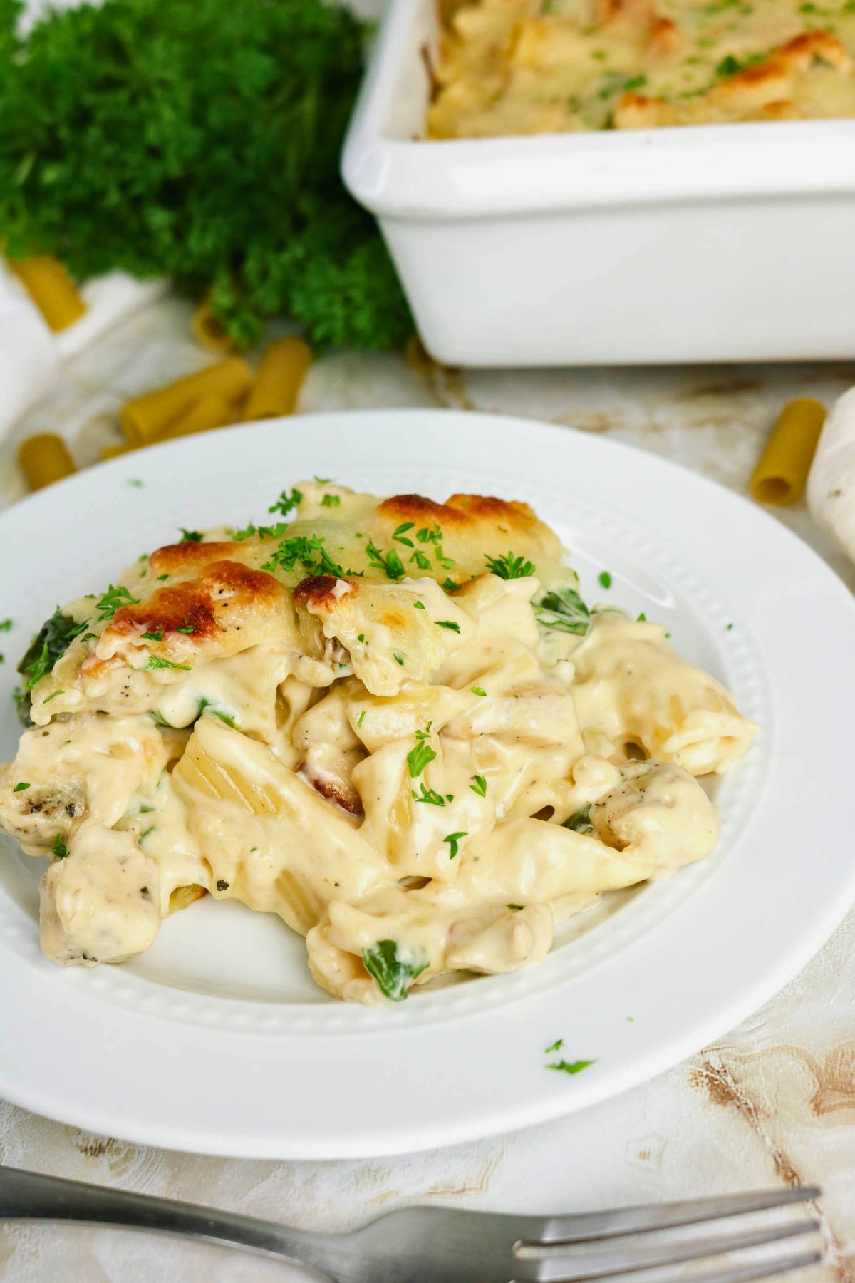 A serving of creamy baked pasta with chicken and herbs on a white plate, with a casserole dish and uncooked pasta in the background.