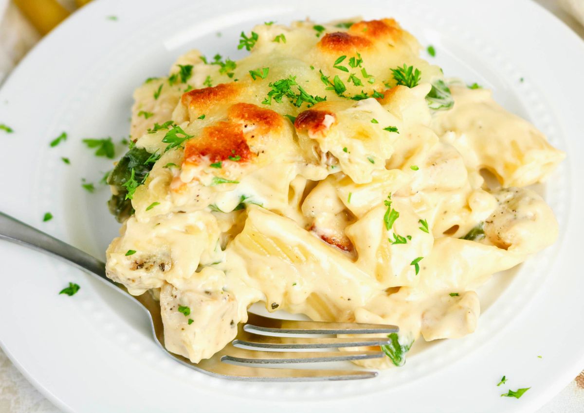 A serving of creamy baked pasta with chicken and herbs, topped with melted cheese and garnished with chopped parsley, on a white plate with a fork.