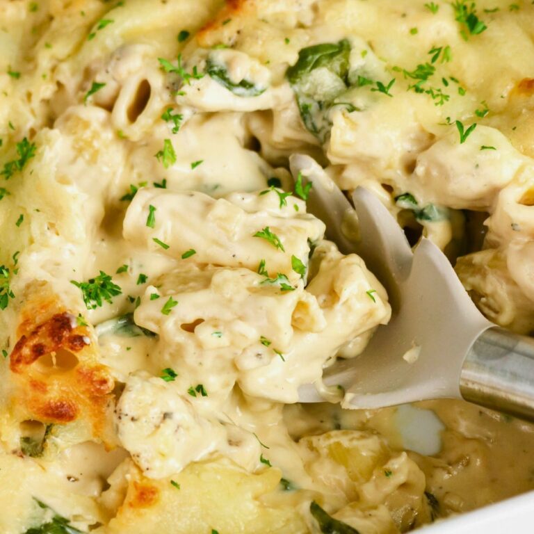 Close-up of creamy baked pasta with chicken, spinach, melted cheese, and herbs, being served with a gray spoon.