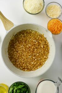 A white pan with sautéed diced onions sits surrounded by bowls of shredded cheese, spinach leaves, milk, and oil on a white surface.