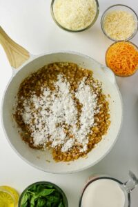 A white pan with sautéed onions and flour, surrounded by bowls of shredded cheese, spinach, milk, and oil on a white surface.