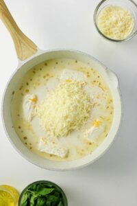 A white skillet filled with a creamy sauce, shredded cheese, and cream cheese blocks. Surrounding bowls contain shredded cheese, spinach leaves, and olive oil.