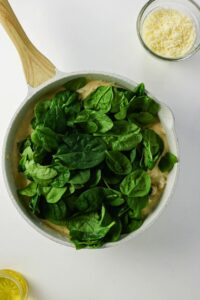 A white skillet filled with creamy sauce and topped with fresh spinach leaves, next to a bowl of grated cheese and a partially visible lemon slice on a white surface.