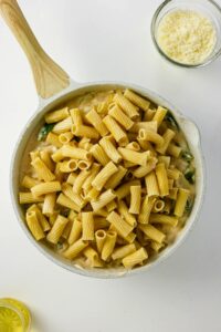 A white skillet filled with uncooked rigatoni pasta on top of creamy sauce, beside a bowl of grated cheese and a small dish with a lemon slice.