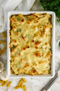 A baked pasta casserole with a golden, melted cheese topping in a white rectangular dish, surrounded by some uncooked pasta, garlic, and fresh herbs on a light surface.