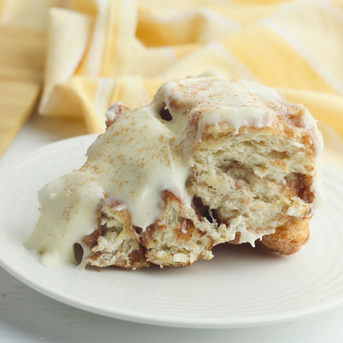 A cinnamon roll topped with cream cheese icing and a sprinkle of cinnamon sits on a white plate with a yellow napkin in the background.