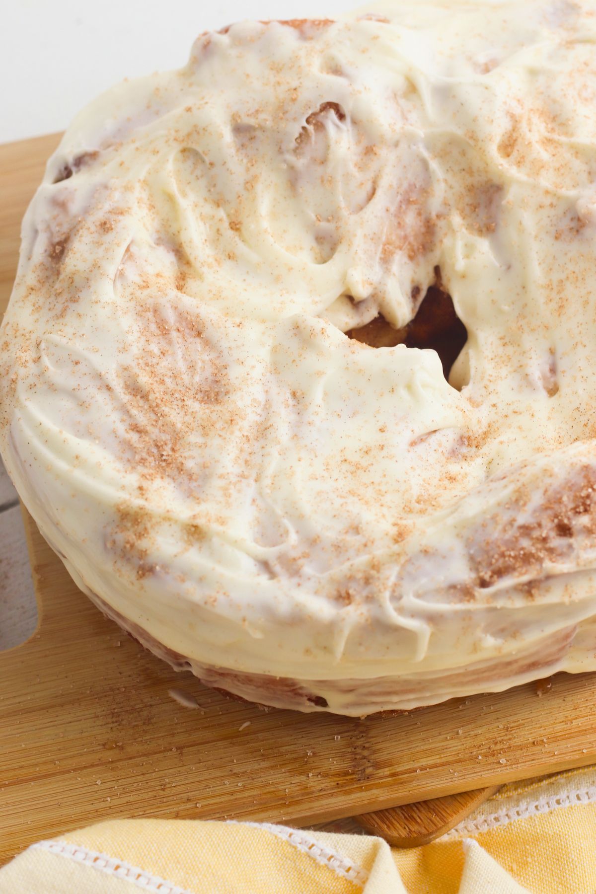 A round cake with white frosting and a dusting of cinnamon sits on a wooden cutting board.