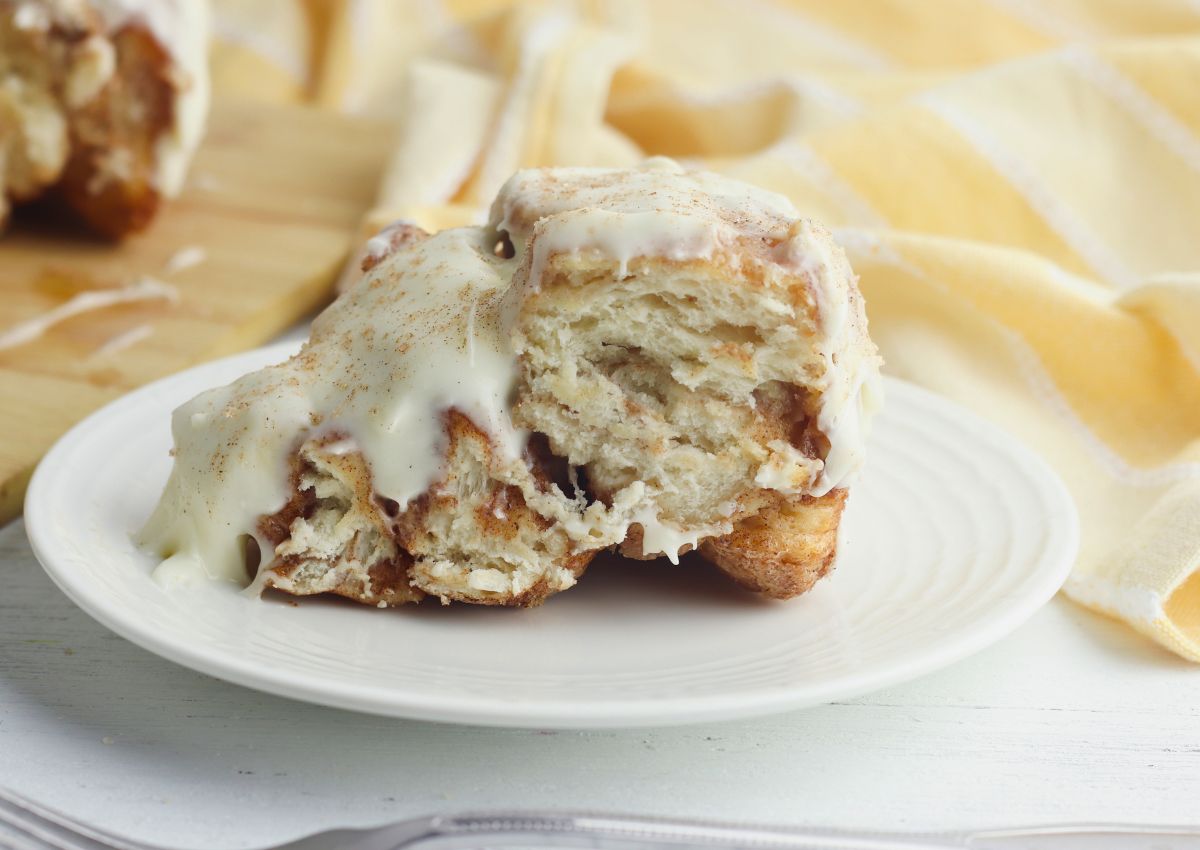 A partially eaten cinnamon roll with white icing on a white plate, set on a white table with a yellow-striped cloth in the background.