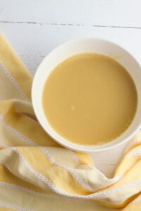A white bowl filled with creamy yellow soup sits on a white surface next to a yellow and white striped cloth.