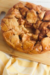 A ring-shaped monkey bread made of cinnamon-coated dough pieces sits on a wooden cutting board next to a yellow-striped cloth.