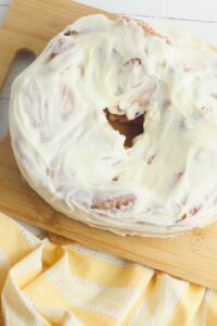 A round cinnamon roll cake with white icing sits on a wooden cutting board next to a yellow striped cloth.
