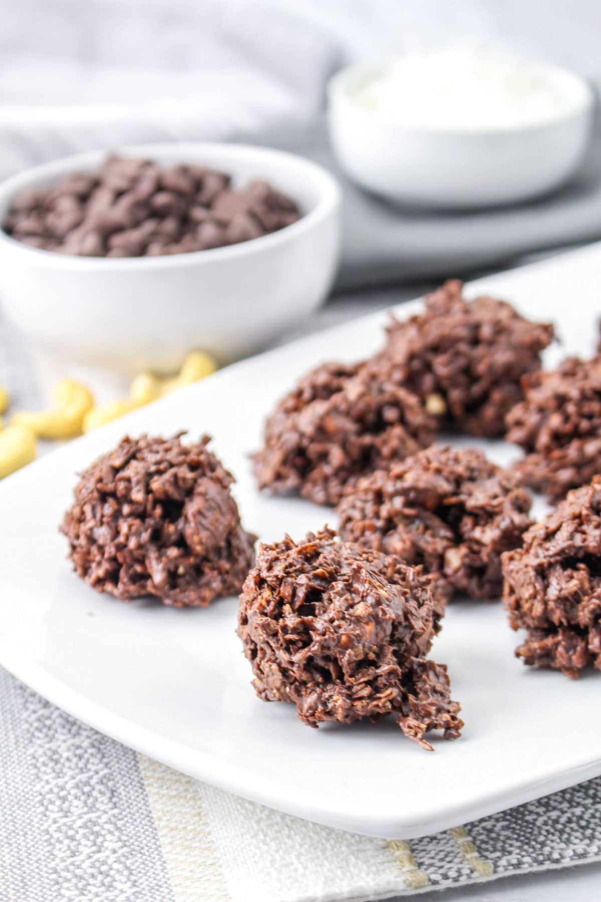 Chocolate no-bake cookies are arranged on a white plate, with a bowl of chocolate chips and another bowl in the background.