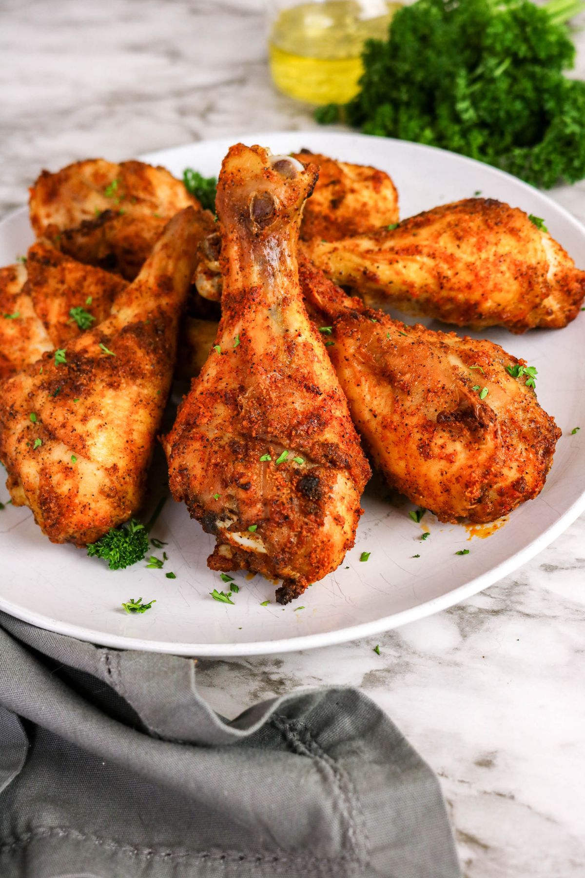 A white plate with several seasoned and cooked chicken drumsticks, garnished with chopped herbs, sits on a marble surface next to a bunch of parsley and a bottle of oil.