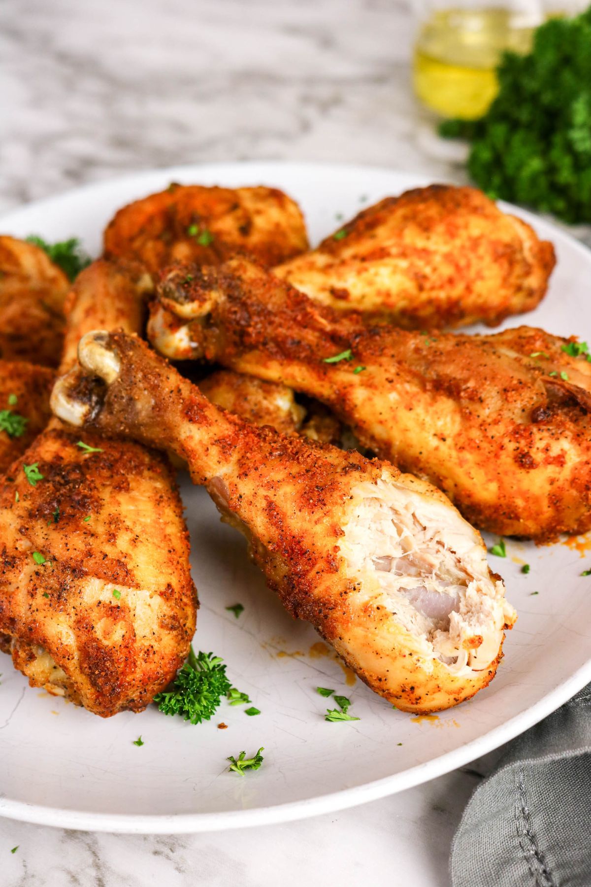 Plate of seasoned, cooked chicken drumsticks garnished with chopped parsley, with one drumstick showing a bite revealing the inside.