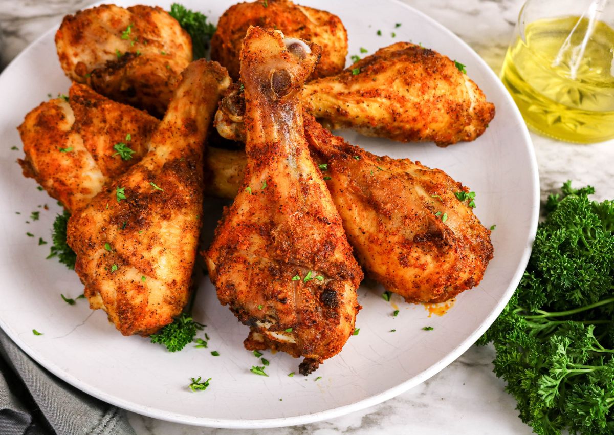 Plate of seasoned, baked chicken drumsticks garnished with chopped parsley, with fresh parsley and a bottle of olive oil nearby on a marble surface.
