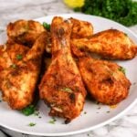 A plate of seasoned, baked chicken drumsticks garnished with chopped parsley, served on a white dish with greens in the background.