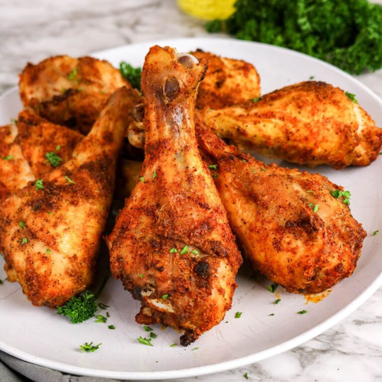 A plate of seasoned, baked chicken drumsticks garnished with chopped parsley, served on a white dish with greens in the background.