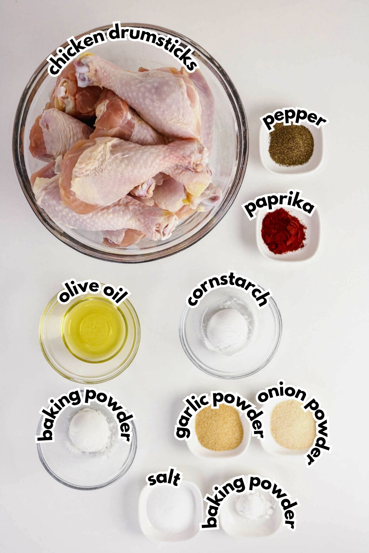 A bowl of raw chicken drumsticks surrounded by small bowls containing pepper, paprika, cornstarch, olive oil, baking powder, garlic powder, onion powder, and salt.