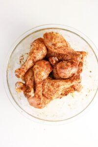 Raw chicken drumsticks coated with seasoning are placed in a clear glass bowl on a white surface.