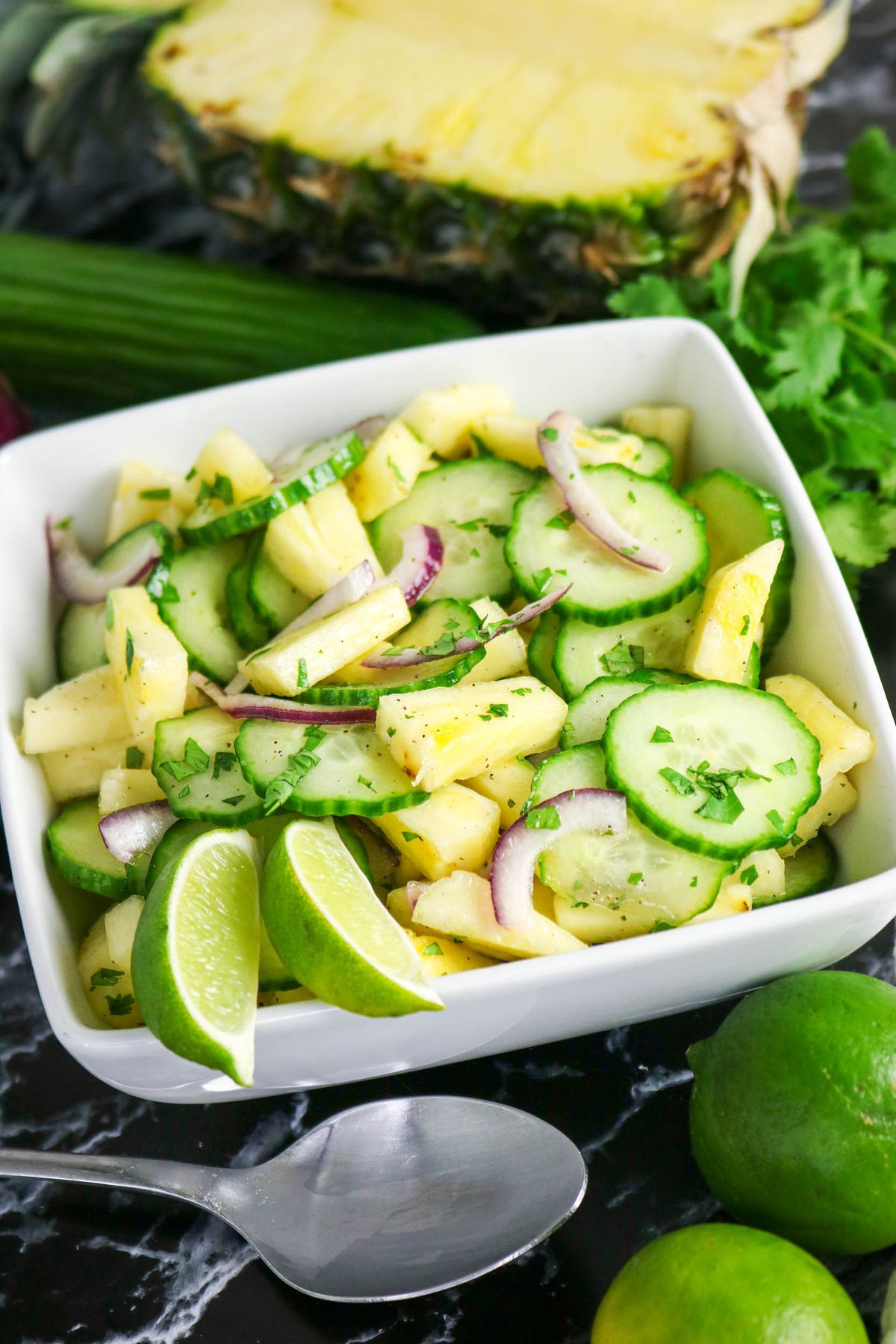 A white bowl filled with a salad of sliced cucumbers, pineapple chunks, red onion, cilantro, and lime wedges, with a spoon and fresh ingredients nearby.