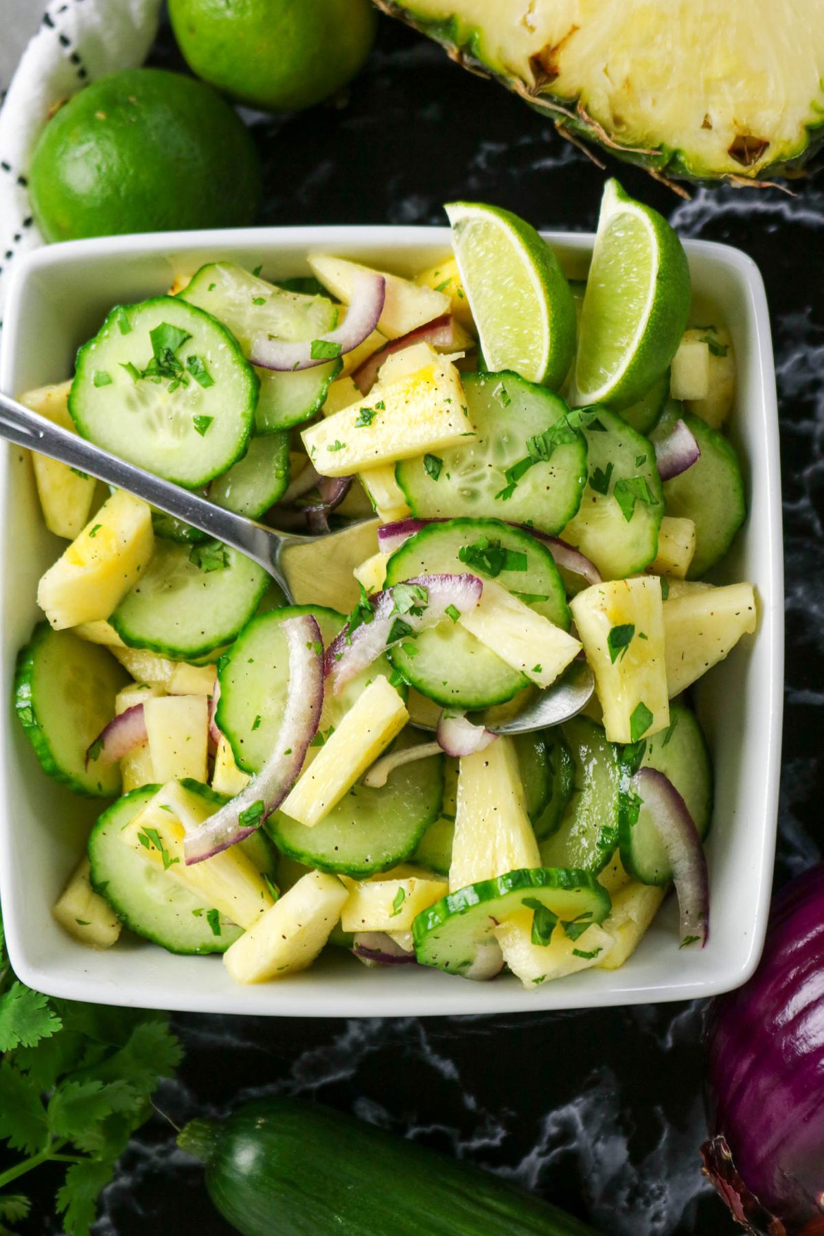 A bowl of cucumber, pineapple, and red onion salad garnished with cilantro and lime wedges, with a spoon resting in the bowl.