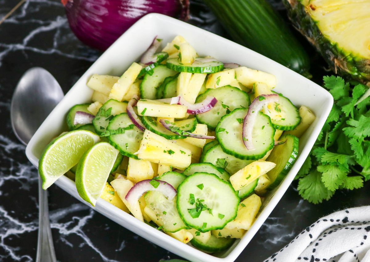 A square white bowl filled with a salad of cucumber, pineapple, red onion, and cilantro, garnished with lime wedges. A spoon, cilantro, and fresh produce surround the bowl.