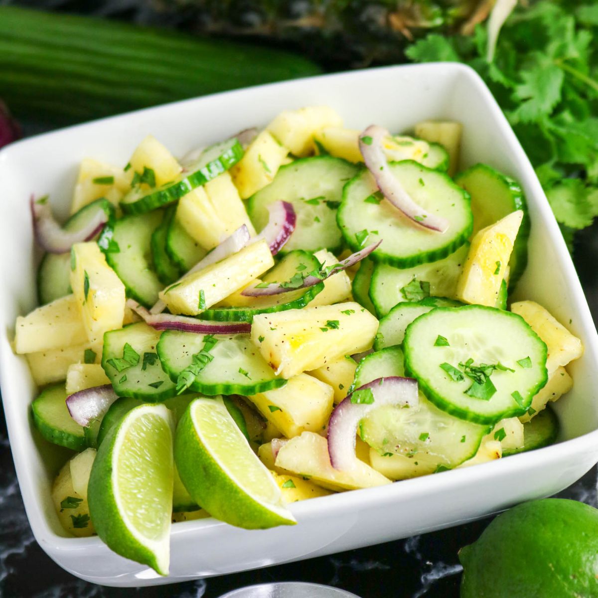 A square white bowl filled with a salad of sliced cucumbers, pineapple, red onion, and chopped herbs, garnished with lime wedges.