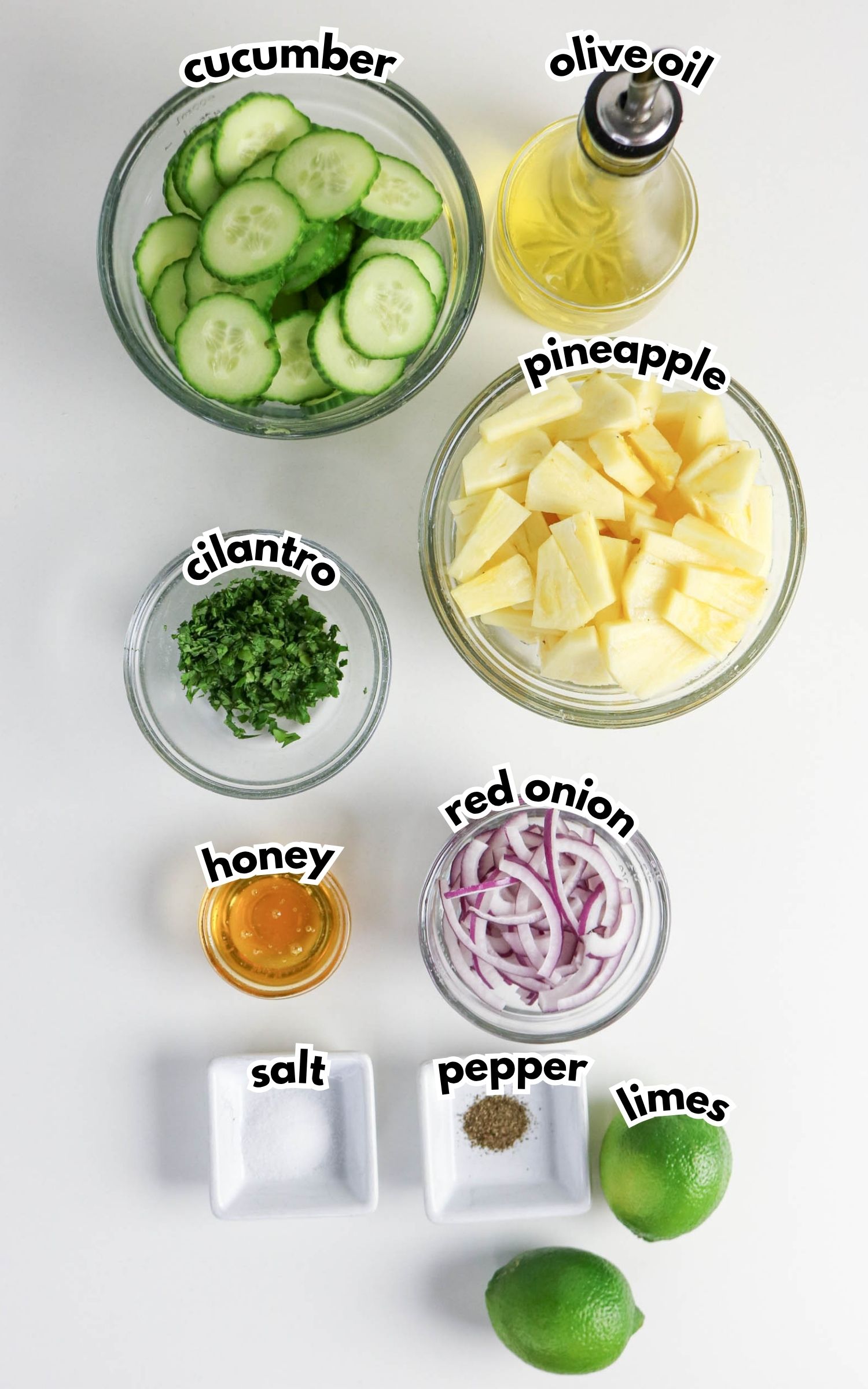 Ingredients for a salad arranged on a white surface: sliced cucumber, olive oil, pineapple chunks, chopped cilantro, honey, red onion, salt, pepper, and whole limes.