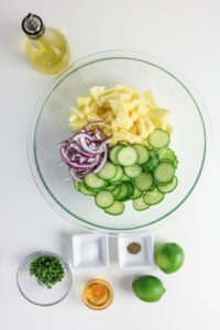 A glass bowl with sliced cucumbers, pineapple, and red onion, surrounded by a bottle of oil, limes, chopped herbs, salt, pepper, and a small bowl of honey.