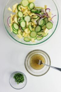 A glass bowl with sliced cucumber, pineapple, and red onion, a small bowl of chopped herbs, and a bowl of vinaigrette with a fork on a white surface.