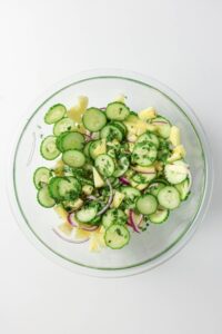 A glass bowl containing a salad made of sliced cucumbers, red onions, potatoes, and herbs on a white background.