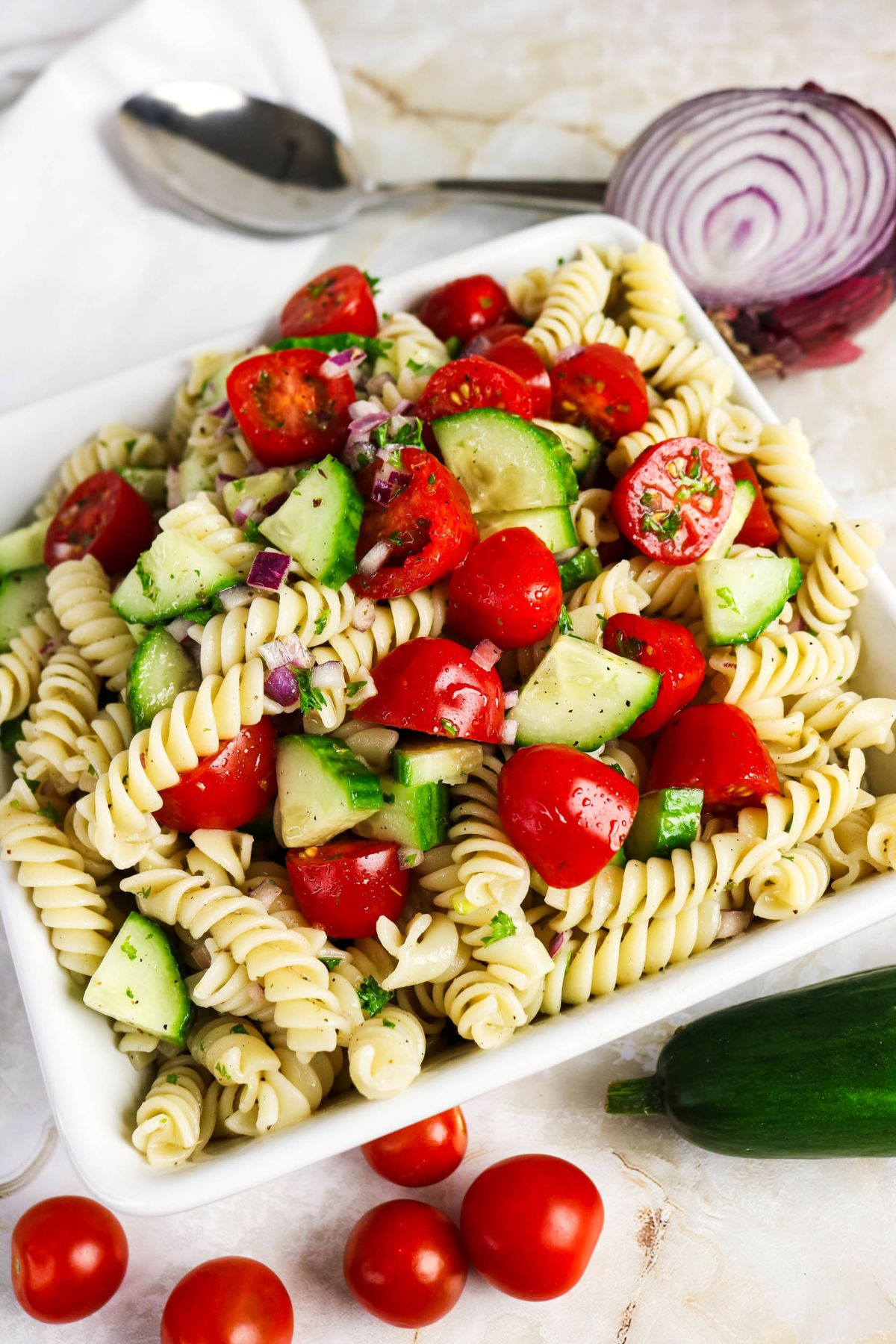 A white dish filled with rotini pasta mixed with chopped cucumbers, cherry tomatoes, red onions, and herbs. Sliced red onion and fresh vegetables are nearby.