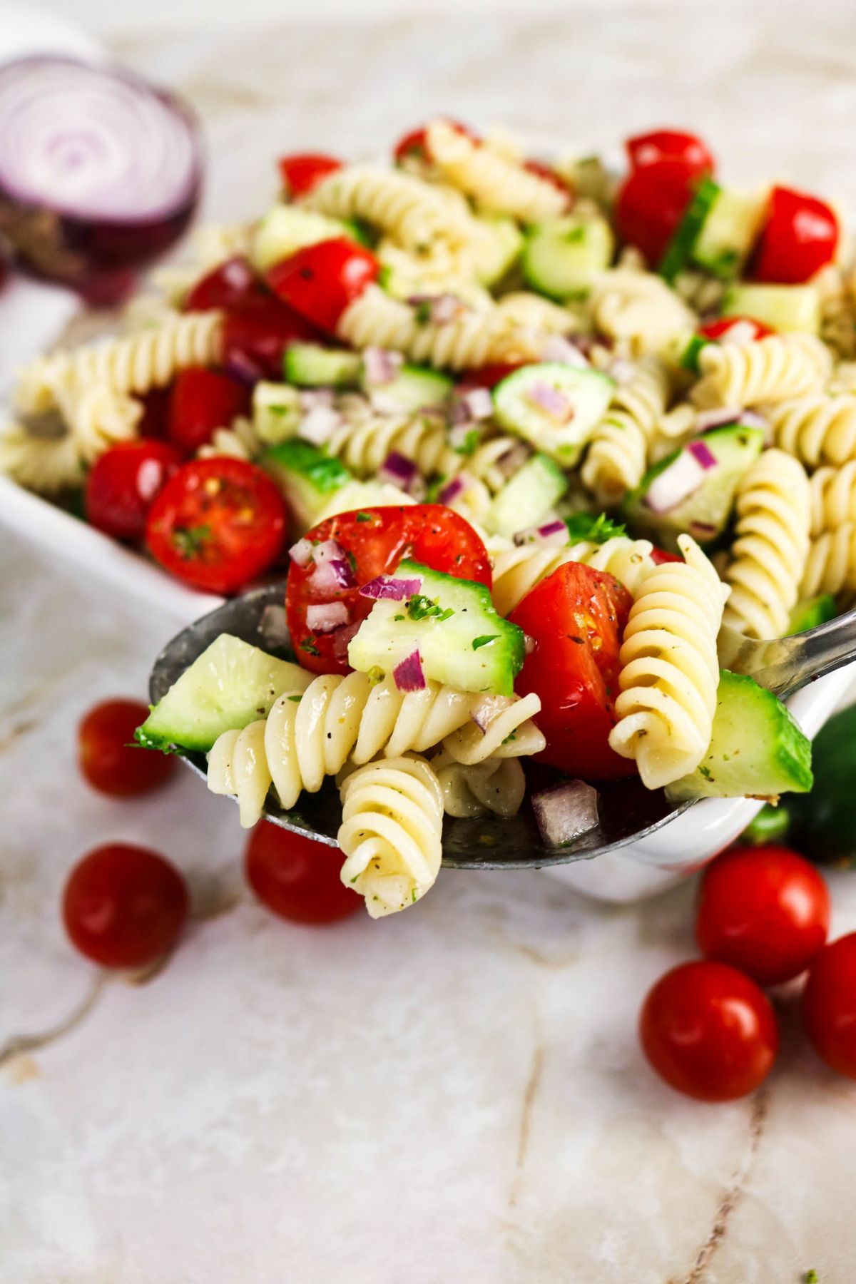 A close-up of a pasta salad with rotini, cherry tomatoes, cucumber, and red onion in a spoon, with a bowl of the salad and loose tomatoes in the background.